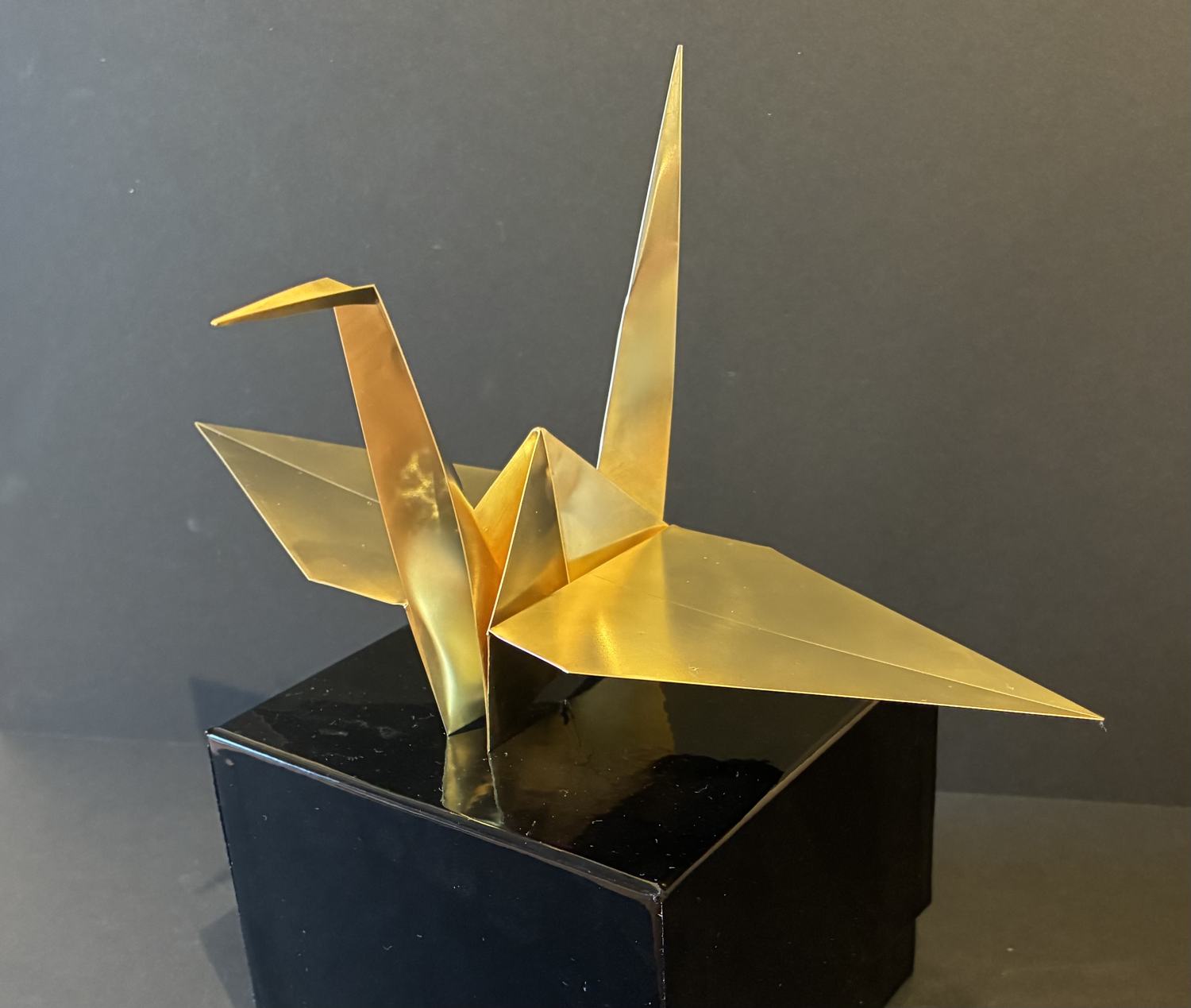 A folded brass origami crane, gold and mirror-bright, standing on a glossy black plinth