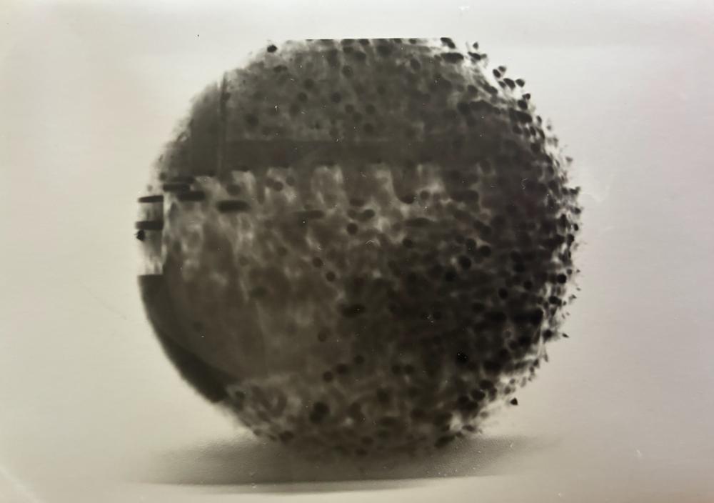 Pinhole photograph: a large dark speckled sphere resting on a pale surface