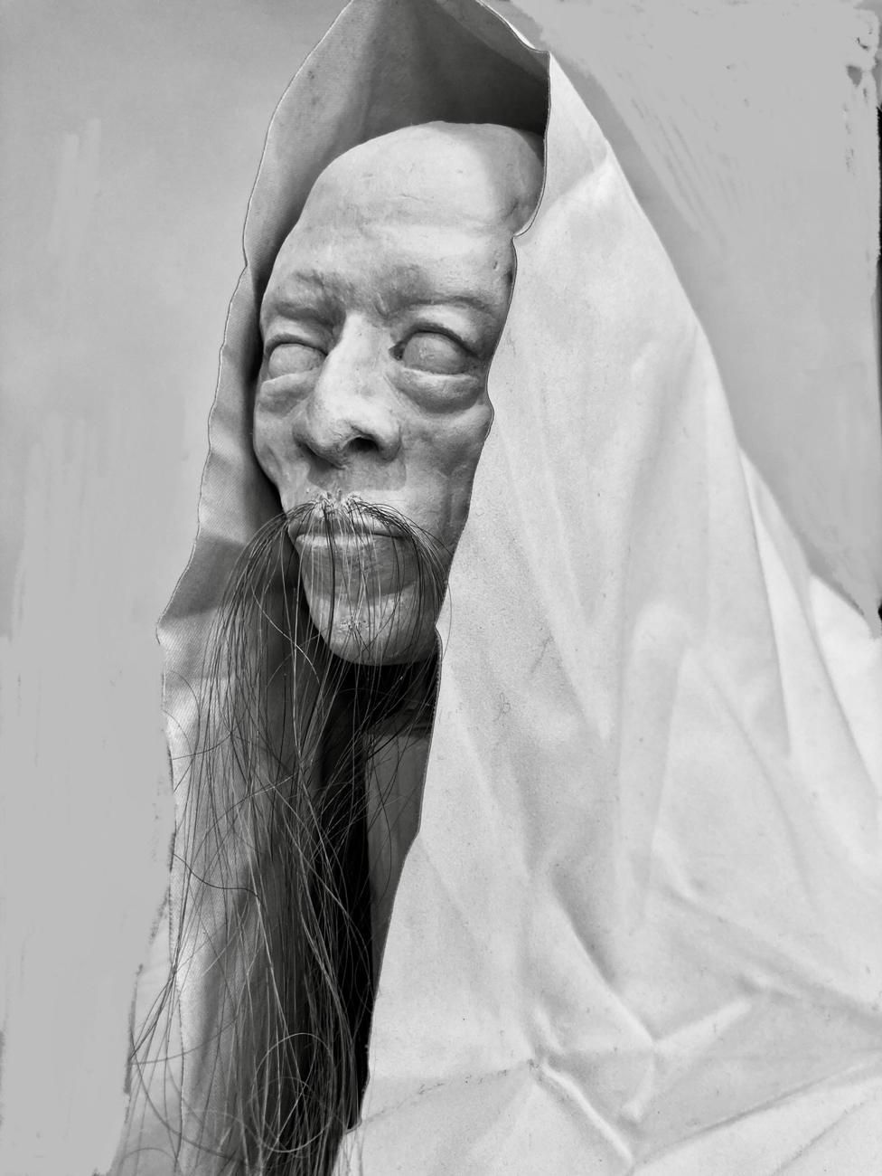 Sculpted head with closed eyes and a long beard of real hair, draped in a pale hood